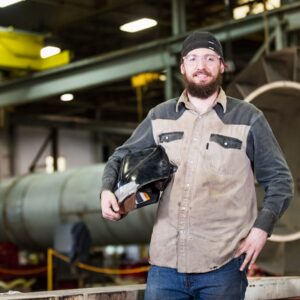 a man in a metal shop holding a welding hat in a welding jacket and safety gear